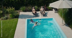 The family relaxes by their pool in the backyard of the house. Adults sunbathe on sun loungers, two children swim in the pool
