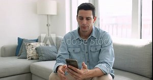 Young serious Hispanic man holding smartphone seated on couch, looks at modern gadget screen, read notification or message feels thoughtful, ponder over answer, consider received information by e-mail