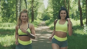 Beautiful women running in park. Two young females jogging outdoors in green sportswear. Blonde and brunette girls running in slow motion. Healthy lifestyle concept. Stabilized shot