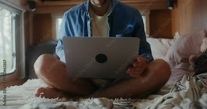 A young man with glasses opens a laptop and starts using the laptop while sitting on a bed in a van