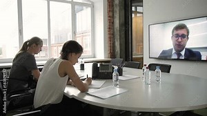 In conference hall there is a videoconference with a business man. Two women sit in front of monitor and record information that speaker says on webinar.