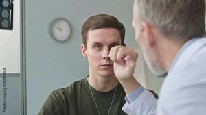 Mid aged military doctor using torch pen with flashlight to check eyesight of young soldier having medical checkup appointment