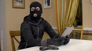 A robber is trying to hack into a tablet. A masked thug is sitting in a house and cannot hack into a tablet. Theft of data from a tablet.