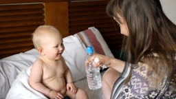 Joyful baby and mother have fun on bed. Mom gives baby bottle of...