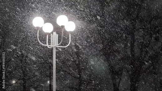 Bright street lamp illuminates heavy snowfall during cold winter night. Snowstorm creates beautiful shimmering effect around light source in dark park.