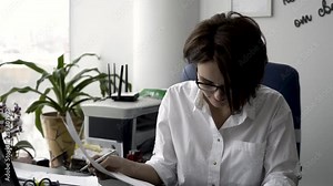 Attractive young office woman in white shirt working on the business papers, signing documents near green plant and printer. Female office worker arranging documents and putting her signature on them.