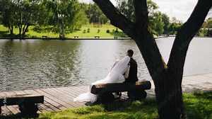 The bride and groom are sitting on a bench by the pond. They look at the water and hug each other.