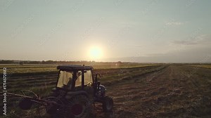Small ranching hay harvesting. Tractor working in field at sunset. Tedder for tractor. The machine for gathering hay.
