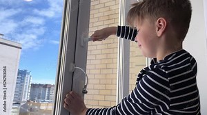 safety at home for small children. a lock on the window protects children from opening the window. The boy tries to open the window standing on the windowsill on a high floor.