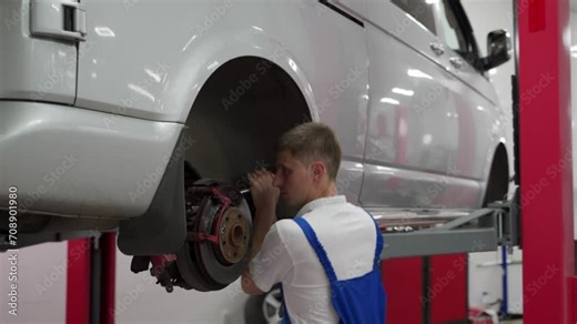 Auto mechanic in uniform fixes vehicle suspension, performs maintenance at car service garage. Male technician inspects shock absorbers, brakes, adjusts wheel assembly alignment