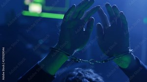 Close-up of hacker�s hands wearing handcuffs. Silly computer hacker caught red-handed and arrested. Dark hideout place with computer equipment in dark office on background.