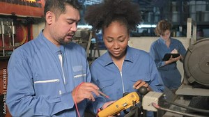 Asian male and female African American engineers in safety uniform work by inspecting machines' voltage current, checking, and maintaining at manufacture factory, electric system service occupations.
