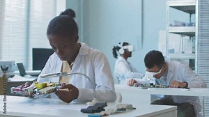 African American female student of computer engineering and her colleagues examining circuit boards of different types in laboratory doing research