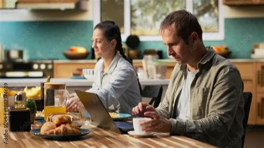 Busy working dad typing on laptop at kitchen table with his family, ignoring his pre-teen girl and wife during breakfast. Distracted preoccupied father distant with his daughter. Camera B.