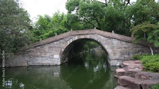 Historic stone arch bridge in Moon Lake Park in Ningbo, China. Traditional bridge crosses a calm canal surrounded by lush trees and classical Chinese garden. Peaceful atmosphere of a historic park