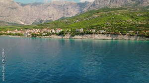 Serene coastal town nestled against a rugged mountain backdrop. Buildings cascade down to meet the blue sea, as waves gently lap rocky shore.