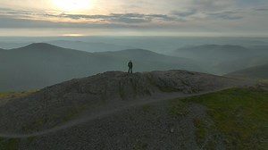 Mountain walker lone figure on fell summit with orbit revealing misty valleys and rounded fell peaks at golden hour | Premium Stock Video Footage