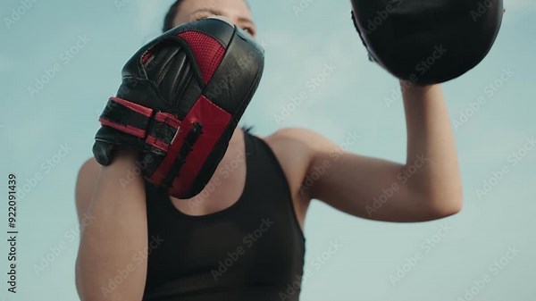 Two women engaging in a boxing workout outdoors with one holding boxing pads for the other in a close-up shot.