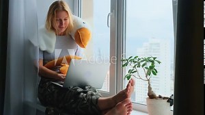 A happy barefoot woman using a laptop while sitting on a windowsill with a funny plush toy. Remote work, blogging, freelance.