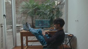 Male programmer sitting with feet up on desk with computer and laptop and texting on phone during break in comfy home office with window to backyard garden. Back view, zoom-out shot