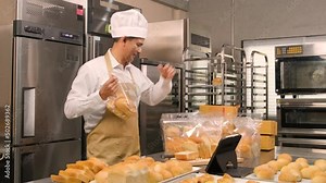 Senior Asian male chef in apron, small business blogger live streaming via mobile application technology, bakery sales online promotion, fresh bread, pastry food, and commercial cooking in kitchen.