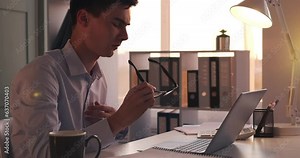 Focused businessman works on laptop. His fingers move across the keyboard, showcasing efficiency and dedication to the task at hand. Amidst work, he takes a moment to clean his glasses.