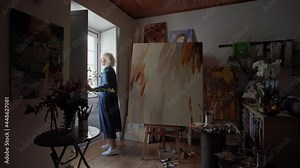 A middle aged woman artist in a dark room of the gallery opens a window. The sun illuminates the art workshop. Paintings painted with acrylic paints. Slow motion