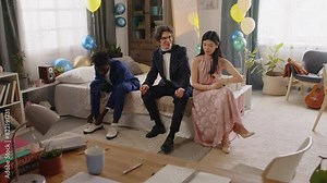 Group of youthful intercultural graduates in suits and evening gown sitting on double bed in domestic room and getting ready for prom party while African American guy tying shoelaces