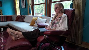 Unhappy frustrated woman sits in a red leather recliner using a laptop and reads bad news with expression. The room is decorated with a white couch and colorful pillows