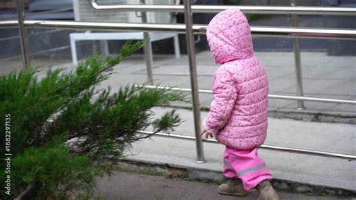 Adorable little kid wearing warm pink puffer coat walks away on sidewalk, moving quickly past parked bikes and disappearing behind dense green shrub