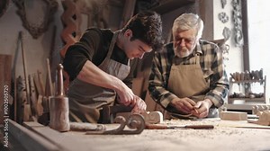 Two carpenters working together. Young man learning how to operate chisel. Older Caucasian man with beard spectating. Showing how everything must be completed. Concept of teaching.