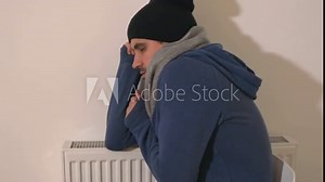 Freezing young man with hoodie, hat and scarf sitting next to a heater and shivering with cold, energy crisis