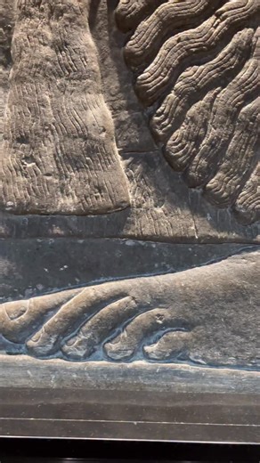 This Assyrian relief shows a winged protective deity performing a ritual act, anointing a sacred object with a perfume cone dipped from a pail he carries. Similar figures, sometimes eagle-headed, appear throughout Assyrian palace art and are thought to symbolize divine protection and purification, often in relation to sacred trees. The figure is richly detailed, with elaborately curled hair and beard, heavy fringed garments, and jewelry, reflecting royal and divine status. An inscription running