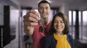 Portrait of young husband and wife holding keys to new home in front of the camera. Happy family smiling and showing keys to new apartment. Slow motion. Keys with wooden house shaped key ring.
