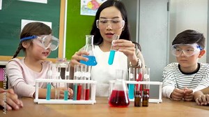 Young woman mixing liquids in test tube with little children watching, learning about science, chemistry. Asian school teacher and students in science class