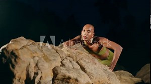 Androgynous black lgbtq fashion model crawls on stones inside picturesque natural pool at night. Non-binary biethnic person poses in still water among rocks in a creek on exotic island. Pride month
