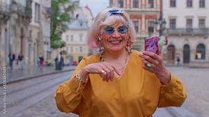 Elderly grandmother woman tourist with smartphone, smiling, listening music in app wearing earphones, dancing, enjoying sightseeing in city center. Travelling vacation. Active life after retirement