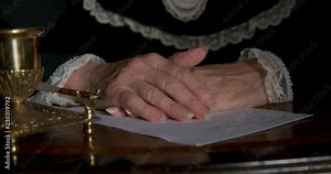 An ornate inkwell with a holder for its fancy dip pen and the folded hands of an old Victorian era woman with an unfinished letter who then exits the scene.