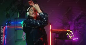 Young joyful beautiful Caucasian woman in black leather coat and headphones dancing in neon room. Pretty happy female dancer in underground bar. Girl in disco. Stock Video