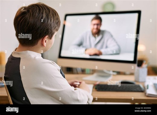 Caucasian boy sitting at desk watching middle aged Caucasian man on computer monitor during video call, boy facing screen with hands resting on table, home interior background visible Stock Photo - Alamy