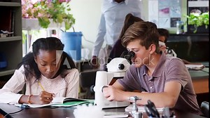 High School Students Looking Through Microscope And Writing Notes In Biology Class