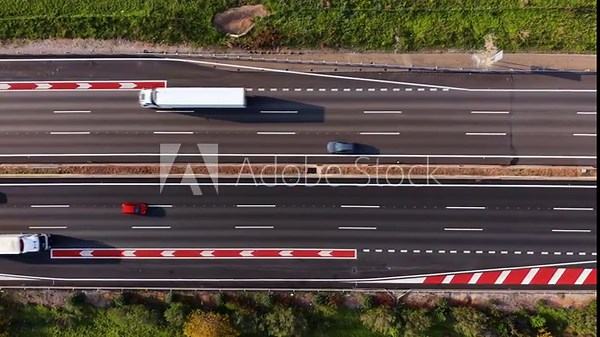 Overhead top-down view of a dual carriageway highway showing multiple lanes of traffic with red and white chevron road markings, white dashed lane lines, trucks and cars in motion