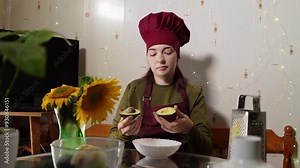 Lady in red chef hat and apron is dividing ripe avocado in to halves and extracting seed filmed in slow motion. Caucasian woman is preparing healthy green product sitting at the table in cozy kitchen.