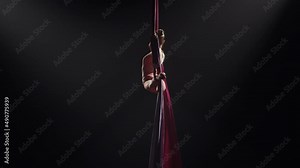 Young female circus gymnast spins on aerial silk and demonstrates stretching. Difficult acrobatic stunts at height on black background with backlight. Video for sports, acrobatic, circus school. Slow.