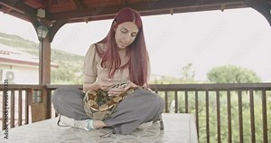 A young woman with red hair sits on a patio table, gently interacting with her pet snake.