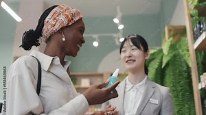 Young black woman smelling new refreshening body mist in cosmetic supermarket while Asian female shop assistant consulting her and helping choose new scented bodycare product