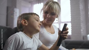 Cute son with TV remote can't decide what television program to watch together with mom. Family spending leisure time at home relaxing on sofa in domestic room and watching cartoonsand movies on TV