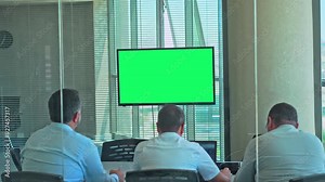 Green screen monitor in a conference room with large windows. View from the back through the glass walls on a company meeting during the day. Blinds on a transparent glass wall