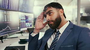 A businessman in a suit looking concerned while sitting in front of computer screens showing charts and data in an office. | Premium Stock Video Footage