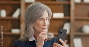 Portrait of female librarian using mobile phone for scrolling social media during work break. Mature woman enjoying reading news while standing near bookshelf at library.